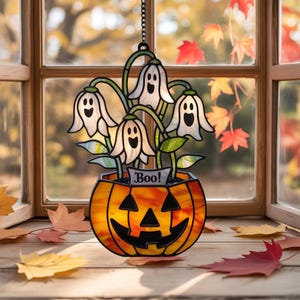 May include: A stained glass Halloween decoration. It features a pumpkin-shaped pot with ghost-like flowers. The flowers are white with black faces and green stems. The pot has an orange and yellow glow, and the word "Boo!" is on a banner.