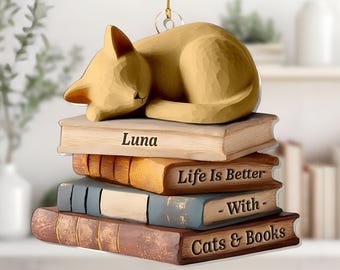 May include: A tan-colored cat ornament sleeping atop a stack of books. The books are various colors and have text that reads "Luna", "Life Is Better - With - Cats & Books". The ornament hangs from a gold string.