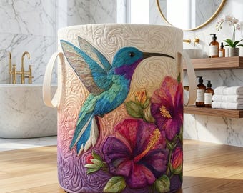 May include: A cylindrical laundry hamper with a hummingbird and hibiscus flower design. The bird is blue, green, and purple, with a long black beak. The flowers are purple and red. The hamper has cream-colored handles.
