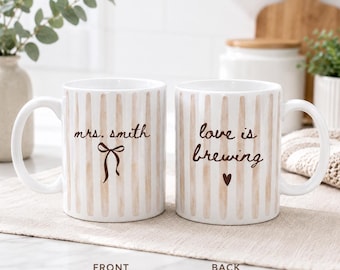 May include: Two white ceramic mugs featuring vertical tan stripes. The left mug displays the text "mrs. smith" above a brown bow graphic. The right mug features the text "love is brewing" with a small brown heart icon below the lettering.