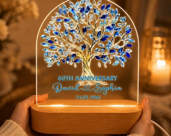 May include: A lighted tree-shaped anniversary gift with blue crystal leaves and a gold trunk. The base is wood, and the text reads "60th Anniversary" with names and a date. The light is held by a person.