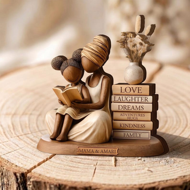 May include: A wooden figurine depicts a mother and child reading a book. The figurine is light brown with a stack of books labeled "Love, Laughter, Dreams, Adventures, Hugs, Kindness, Family." The base reads "Mama & Amari."