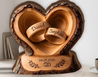 May include: A heart-shaped wooden ring holder with natural bark edges. Two wooden rings are inside, engraved with the names David and Sophia. The base is engraved with the date 04.05.2026, two interlocking rings, and leaf branch designs.