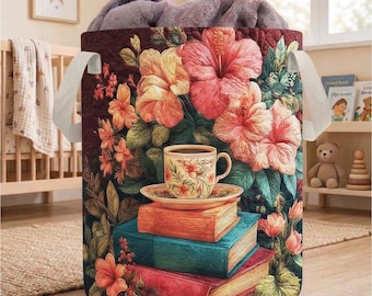 May include: A round laundry hamper with a floral design featuring hibiscus flowers in shades of pink, orange, and yellow. A stack of books and a teacup and saucer are in front of the hamper. The hamper has white handles and is filled with a gray blanket.