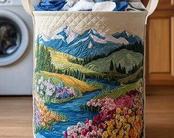May include: A round laundry hamper with a quilted landscape design featuring mountains, a river, and colorful flowers. The hamper is filled with folded towels and has cream-colored handles.