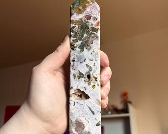 May include: A polished stone tower with a hexagonal point, featuring a complex pattern of white, cream, moss green, and earthy brown inclusions. The mineral surface displays natural crystalline vugs and varied textures throughout its length.