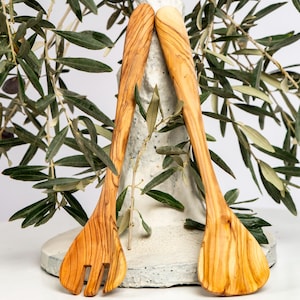 Può includere: Un paio di posate da insalata in legno chiaro, con un'estremità a forma di forchetta e l'altra a forma di cucchiaio. Sono esposte davanti a rami di ulivo con foglie verdi e olive.