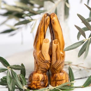 May include: A wooden sculpture of two hands clasped in prayer. The hands are carved from olive wood, showcasing the natural grain and color variations. Green olive branches frame the sculpture against a white background, creating a serene and spiritual aesthetic.