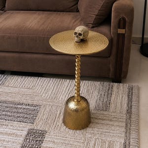 May include: A gold-colored metal side table with a circular top featuring a textured, dimpled surface, a central twisted pedestal, and a wide, hammered bell-shaped base. A realistic, off-white human skull replica sits on the table surface.