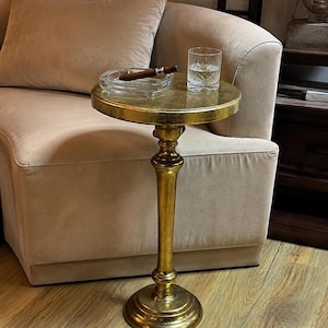 Puede incluir: Una mesa auxiliar alta y redonda de metal dorado con una superficie circular lisa y una base pesada. Sobre la mesa hay un cenicero de cristal transparente con un cigarro marrón y un vaso de cristal tallado con una bebida líquida.