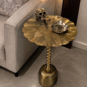 Op de afbeelding: Een goudkleurige metalen bijzettafel met een rond blad voorzien van een textuur met bloemblaadjespatroon, een gedraaide centrale kolom en een afgeronde voet. Op de tafel staan een metalen menselijke schedel met een sigaar en een zilverkleurige asbak.