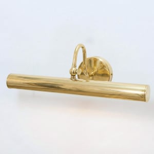 May include: A brass picture light. The light features a cylindrical body, a curved arm, and a circular base. The polished brass fixture is designed for illuminating artwork. It is mounted on a white wall. The light's length is approximately 30 cm.