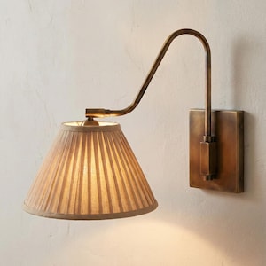 Op de afbeelding: Een wandlamp met een geplooide lampenkap. De armatuur heeft een messing afwerking, met een gebogen arm die zich uitstrekt vanaf een rechthoekige achterplaat. De lampenkap is beige en kegelvormig en geeft een warme gloed.