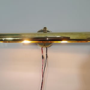 May include: A brass picture light with a cylindrical shade, mounted on a circular base. The light is illuminated, casting a warm glow. Exposed red and black wires are visible. The fixture is designed to illuminate artwork.