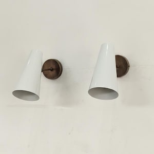May include: Two white cone-shaped wall sconces with bronze-coloured circular backplates. The sconces are mounted on a white wall. The lamps have a mid-century modern design.