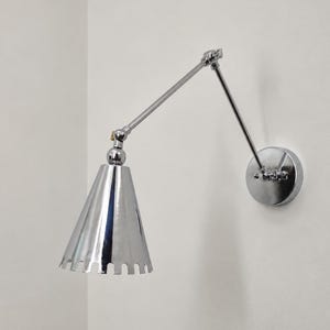 Peut inclure: Une applique murale en chrome poli dotée d'un bras articulé réglable et d'un abat-jour conique avec un bord inférieur festonné. Le luminaire est fixé sur une plaque de base circulaire en chrome, présentant une finition métallique réfléchissante.