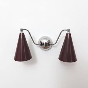 May include: A wall-mounted light fixture with two maroon cone-shaped shades. The shades are attached to a chrome fixture with a central round base. The fixture is mounted on a white wall.