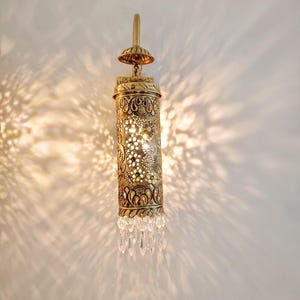 May include: A cylindrical brass wall sconce featuring intricate laser-cut floral and geometric patterns that cast light patterns on the wall. The fixture is topped with a small dome and finished at the bottom with a row of clear glass crystals.