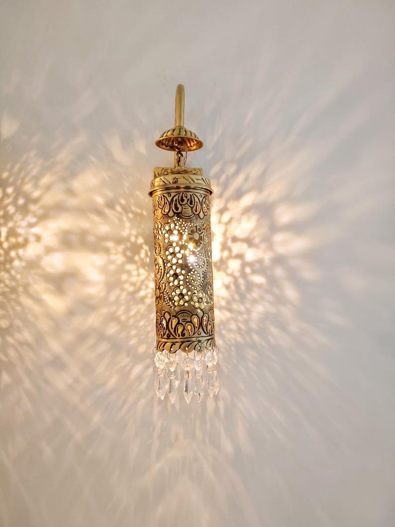 May include: A cylindrical brass wall sconce featuring intricate laser-cut floral and geometric patterns that cast light patterns on the wall. The fixture is topped with a small dome and finished at the bottom with a row of clear glass crystals.