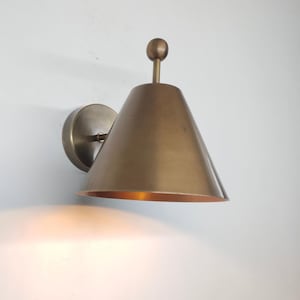 May include: A brass-toned wall sconce with a conical shade and a small, round finial. The sconce is mounted on a circular base and emits a warm, inviting light. The fixture is designed for indoor use and adds a touch of vintage style.