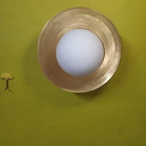 May include: A modern wall sconce with a gold-colored, circular frame and a white, spherical light fixture. The sconce is mounted on a green wall. A small, yellow umbrella-shaped hook is also on the wall.