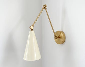 Può includere: Una lampada da parete con un braccio in metallo color ottone dotato di due giunti regolabili e una piastra circolare. La lampada include un paralume conico liscio color bianco sporco che dirige la luce verso il basso.