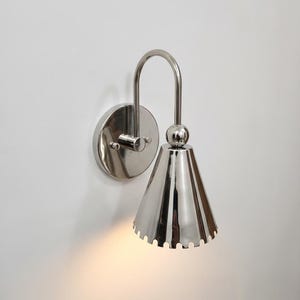 May include: A polished silver-tone wall sconce featuring a circular mounting plate, a curved arm, and a downward-facing conical shade with a scalloped bottom edge. The fixture emits a warm light from the base of the cone, highlighting its reflective metallic surface.