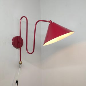 Può includere: Una lampada da parete rossa con un paralume conico. La lampada ha un braccio curvo e una base circolare. Il paralume è angolato verso il basso e la lampada è montata su una parete bianca. Il cavo della lampada è visibile.
