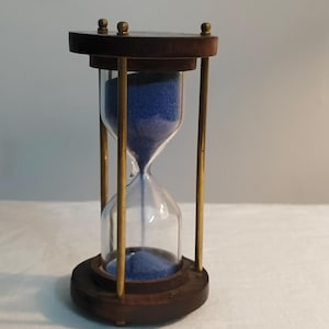May include: An hourglass with a dark wooden frame and brass supports. The glass container holds blue sand. The hourglass has a round base and top, and is a decorative item.