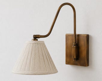 Op de afbeelding: Een wandlamp met een vierkante messing muurplaat en een gebogen arm die een conische, geplooide lampenkap van gebroken wit textiel ondersteunt. De metalen onderdelen hebben een geborstelde antiek messing afwerking en de kap heeft een textuur.