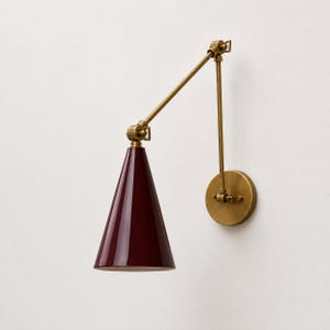 Puede incluir: Una lámpara de pared con una pantalla cónica de color burdeos intenso y un brazo articulado de latón. El aplique incluye una base circular de latón y juntas móviles que permiten ajustar la posición de la fuente de luz.
