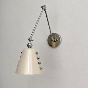 Puede incluir: Una lámpara de pared con una pantalla cónica de color crema adornada con pequeños tachones esféricos de tono plateado. El aplique incluye un brazo ajustable de cromo pulido y una placa de pared circular, ofreciendo iluminación funcional.