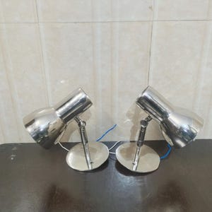 Könnte beinhalten: Zwei polierte Chrom-Schreibtischlampen mit verstellbaren Köpfen und kreisförmigen Sockeln. Jede Lampe hat einen zylindrischen Körper und einen kleinen, runden Sockel. Die Lampen sind mit blauen und weißen Drähten verbunden. Die Lampen stehen auf einer dunklen Oberfläche.