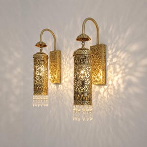 May include: Two wall-mounted sconces featuring cylindrical brass-toned metal shades with intricate cut-out patterns. Each fixture has a curved arm, a rectangular backplate with embossed floral details, and a row of clear crystal-like glass drops.