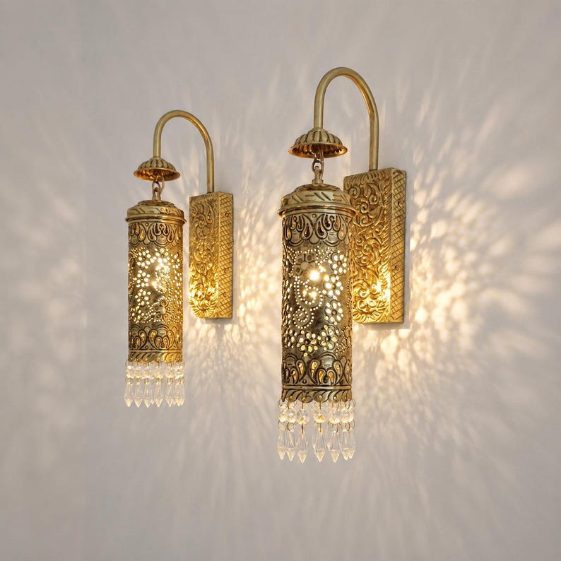 May include: Two wall-mounted sconces featuring cylindrical brass-toned metal shades with intricate cut-out patterns. Each fixture has a curved arm, a rectangular backplate with embossed floral details, and a row of clear crystal-like glass drops.