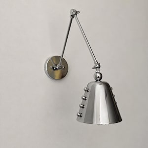 May include: A polished chrome wall lamp featuring an adjustable articulated arm and a conical shade. The shade is accented with a vertical row of four small spherical studs on its side, connected to a circular wall-mounted base plate.