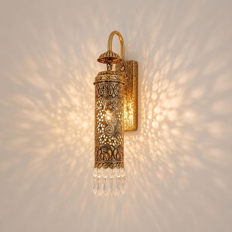 May include: A wall-mounted light fixture with a cylindrical, antique gold-toned metal shade featuring intricate floral cutouts. The lamp includes a rectangular backplate, a curved arm, and a row of clear, dangling crystal prisms at the base.
