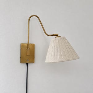 Peut inclure: Une applique murale composée d'une plaque rectangulaire en laiton et d'un bras courbé en laiton. Le luminaire comprend un abat-jour conique en tissu plissé blanc cassé et un cordon d'alimentation noir sortant de la base de fixation.