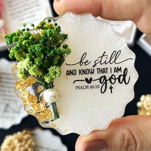 Puede incluir: Un posavasos redondo blanco de resina con un diseño de hoja de oro y un ramo de flores secas verdes. El posavasos tiene el texto "be still and know that I am god" con el versículo "PSALM 46:10" y un símbolo de corazón.