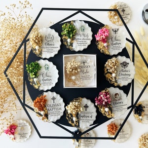 May include: A collection of personalized baptism and first communion favors. Each favor is a unique, scalloped-edged coaster with dried flowers, gold leaf accents, and a pearl. The favors feature names and dates.