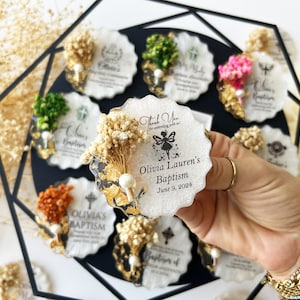 May include: A collection of decorative baptism favors. Each favor is a white, scalloped-edge coaster with dried flowers, gold leaf accents, and personalized text. The favors are arranged on a black surface, with one held in a hand.