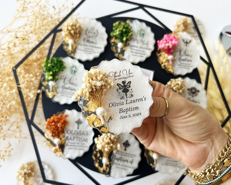 May include: A collection of decorative baptism favors. Each favor is a white, scalloped-edge coaster with dried flowers, gold leaf accents, and personalized text. The favors are arranged on a black surface, with one held in a hand.