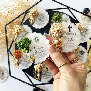 May include: A collection of personalized baptism favors. Each favor is a white, scalloped-edge resin piece, adorned with dried flowers, gold accents, and a cross. The favors are inscribed with "Olivia's Baptism" and the date, June 17th, 2024.