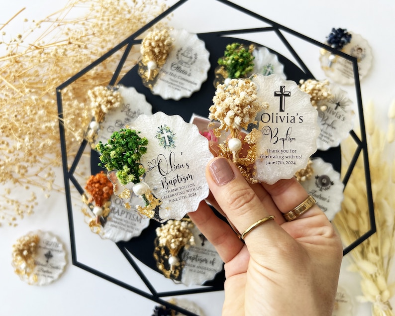 May include: A collection of personalized baptism favors. Each favor is a white, scalloped-edge resin piece, adorned with dried flowers, gold accents, and a cross. The favors are inscribed with "Olivia's Baptism" and the date, June 17th, 2024.