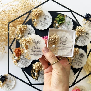May include: A collection of personalized favors for religious events. Each favor is a unique, irregularly shaped resin piece, adorned with dried flowers, gold leaf, and a pearl. Text includes names and dates, perfect for baptisms and confirmations.