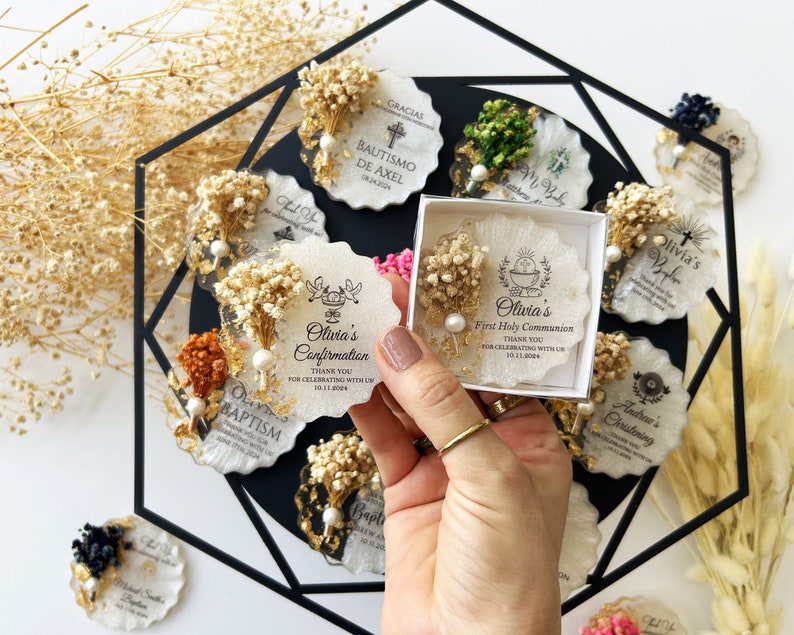 May include: A collection of personalized favors for religious events. Each favor is a unique, irregularly shaped resin piece, adorned with dried flowers, gold leaf, and a pearl. Text includes names and dates, perfect for baptisms and confirmations.