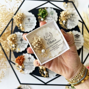May include: A box of First Holy Communion favors, featuring a white resin design with dried flowers, a pearl, and gold accents. The box contains a favor with the text "Olivia's First Holy Communion THANK YOU FOR CELEBRATING WITH US 10.11.2024". Other favors are visible in the background.