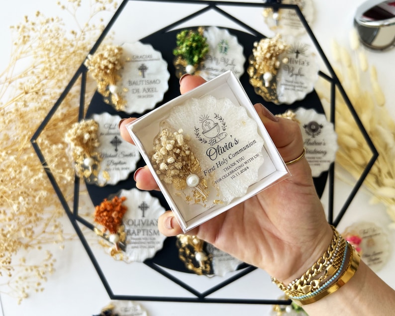 May include: A box of First Holy Communion favors, featuring a white resin design with dried flowers, a pearl, and gold accents. The box contains a favor with the text "Olivia's First Holy Communion THANK YOU FOR CELEBRATING WITH US 10.11.2024". Other favors are visible in the background.