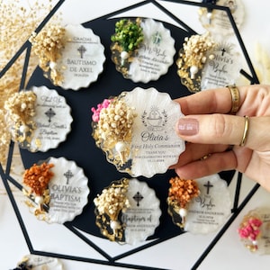 May include: A collection of personalized, scalloped-edge favors, each featuring dried flowers, gold accents, and event details. The favors are white with gold flecks and include text for baptisms and first communions.