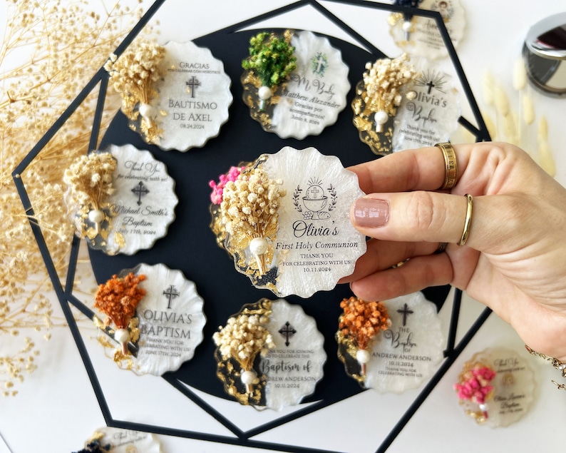 May include: A collection of personalized, scalloped-edge favors, each featuring dried flowers, gold accents, and event details. The favors are white with gold flecks and include text for baptisms and first communions.